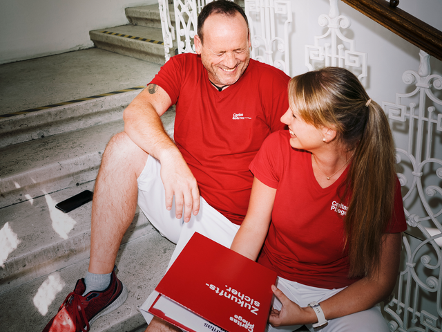 Ein Mann und eine Frau mit roten T-Shirts mit Caritas-Logo sitzen auf den Stiegen und lachen gemeinsam. Die Frau hat einen Aktenordner in der Hand.