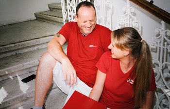 Ein Mann und eine Frau mit roten T-Shirts mit Caritas-Logo sitzen auf den Stiegen und lachen gemeinsam. Die Frau hat einen Aktenordner in der Hand.