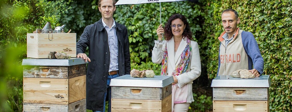 Zwei Männer und eine Frau stehen nebeneinander. Vor ihnen stehen drei Kisten Bienenstöcke.