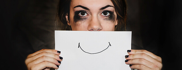 Eine Frau mit dunklen Haaren und schwarz verschmierter Schminke unter den Augen hält ein Blatt Papier mit einem lächelnden Mund vor ihr Gesicht.
