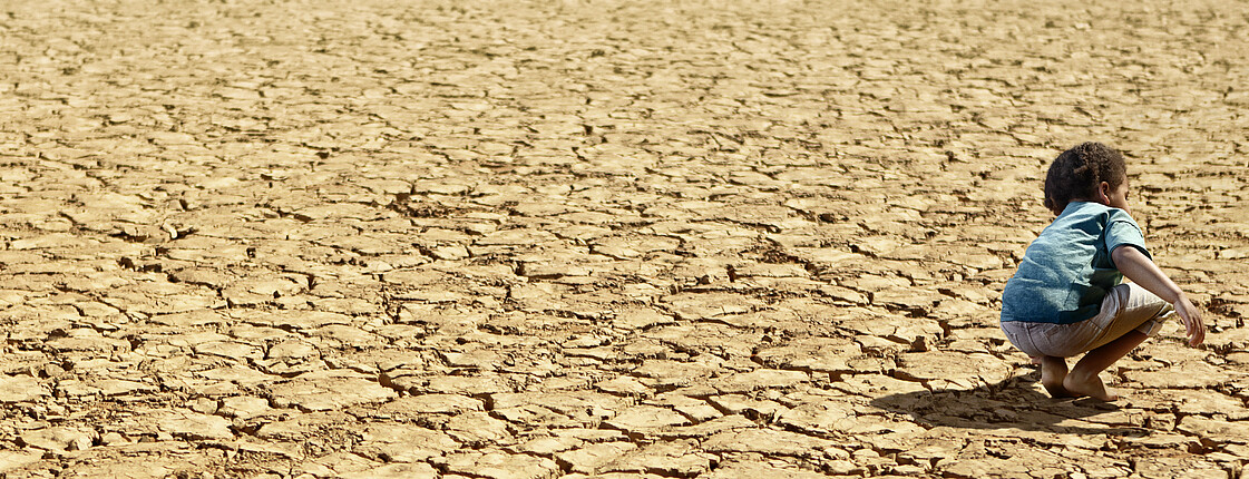 ein kleines Kind hockt barfuß in einer ausgetrockneten Landschaft, welche bis zum Horizont von Dürre gezeichnet ist